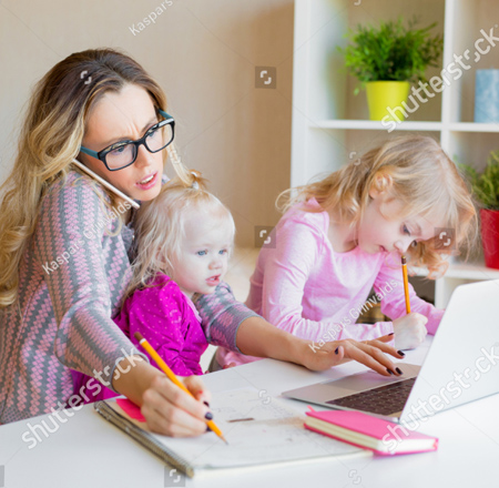 A photo of a woman wearing a pink and grey shirt and glasses. She is holding a pencil in her right hand and is writing on a pad of paper.  She is working on her computer with her right hand.  To her left is a young girl wearing a pink shirt.  She is holing a pencil and writing on a piece of paper.  On the woman's lap is another little girl wearing a pink dress.