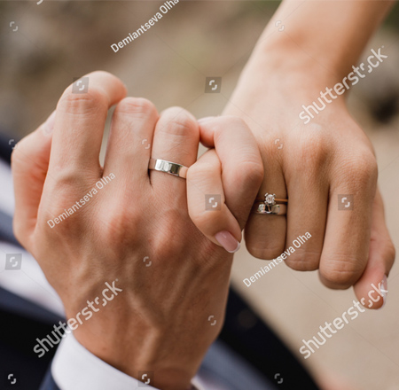 This is a photo of two hands, one male and the other female.  Their pinkie fingers are hooked together.  On the man's hand is a wedding ring. On the woman's hand is both an engagement ring and wedding ring.