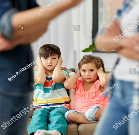 A photo of two children sitting on a couch facing the camera.  The girl on the right is wearing a peach colored shirt and blue pants.  The boy on the left is wearing green pants and a striped shirt.  Both kids look upset and have their hands over their ears. In the foreground, but blurry are the bodies of a an and a woman who appear to be in an argument.