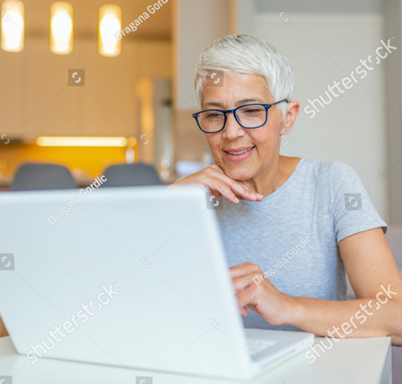 A photo of a woman wearing a grey t-shirt and glasses.  She is sitting at a table working on her laptop computer. She is smiling.