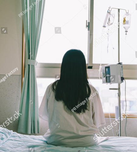 A photo of a woman sitting on a hospital bed looking out the window.  She has her back towards us so we can not see her face.  She has long brown hair and is wearing a hospital gown. There is an IV pole to her right.