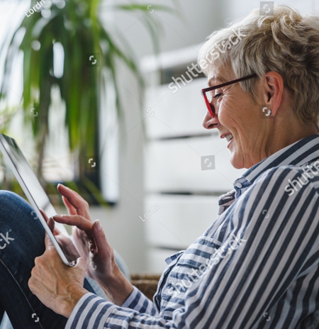 A photo of a woman sitting on a chair.  We see her from the left side and she is looking at her tablet so we don't see her full face.  She is wearing a blue and white striped shirt and blue jeans. She is smiling as she touches the screen of her tablet.