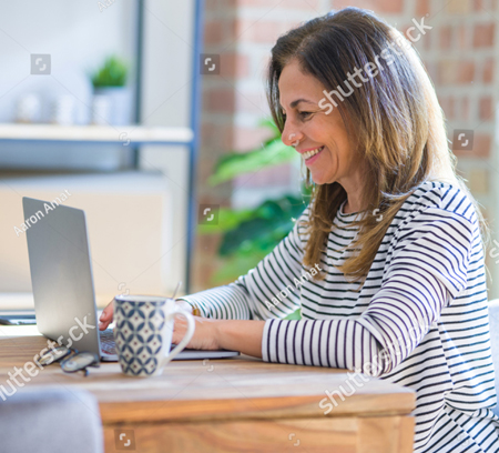 A photo of a woman sitting at a wooden desk working on her laptop computer.  She is wearing a blue and white striped shirt.  She is smiling as she looks down at the screen of her computer.