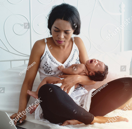 A photo of a woman sitting cross legged on her bed. She is wearing a white top with a black design on it.  In her left arm is an infant dressed in a diaper who is crying. With her right hand she is working on her laptop. The woman has a slightly distressed look on her face.