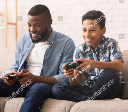 A photo of what appears to be a father and son playing video games.  They are both sitting on a couch, facing us, each with smiles on their faces.  They each have a game controller in their hands. The father is wearing jeans, a white t shirt and a blue shirt over it.  The boy is wearing black jeans and blue plaid shirt.