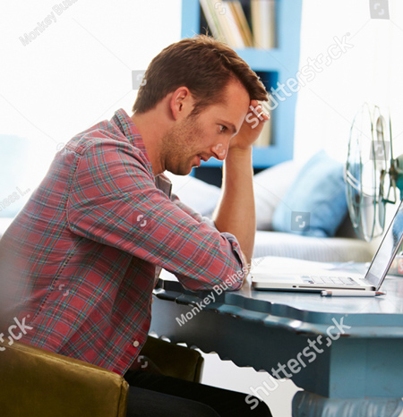 A photo of a man sitting at a blue desk with his laptop computer open in front of him.  He is wearing a red plaid shirt.  The man is leaning forward with his head resting on his right hand.