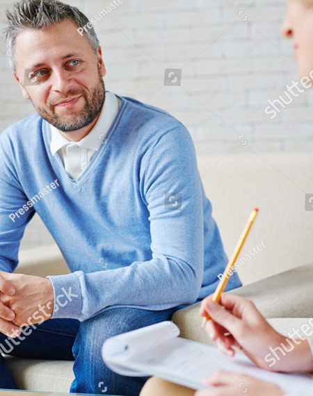 A photo of a man who is wearing blue jeans, a white shirt and a blue sweater.  he is facing us and smiling.  To his left is a blurry image of a woman holding a pencil taking notes.