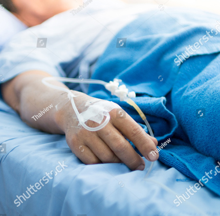 This is a photo of a man laying in a hospital bed.  You see him in a blue hospital gown with blue blankets  over him. The entire image is blurry except for his right hand which has an IV going into it. 