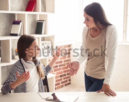 A photo of a girl sitting at a table.  She has her hands up as if to say "what!?" to the woman who is standing to her left.  The girl is wearing a white t shirt and a black and white striped sweater. The woman is standing with her left hand leaning on the table. That woman is wearing blue pants, a beige top with a white sweater over it.  In the background is a bookcase with a few books in it.