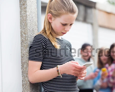 In the foreground of this photo is a young teen girl wearing a black and white stripped shirt.  She is looking down at her phone and has a sad or distressed look on her face. In the back ground, blurry is a group of girls who are also on their phones.