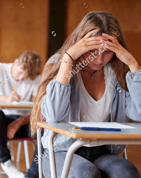 A photo of a teen girl sitting at a desk taking a test.  She is wearing black jeans, a white t shirt with a blue shirt over it.  She is leaning forward with both elbows on the desk in front of her and her head is in her hands. She has a worried expression on her face.  On the desk is a paper and pen. Blurry, in the back ground, is a teen boy taking a test.