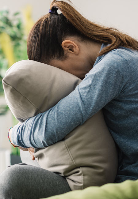 A photo of a woman sitting on a couch.  She is not facing us.  She is wearing grey pants and blue shirt.  She is clutching a grey pillow and her face is buried in the pillow.