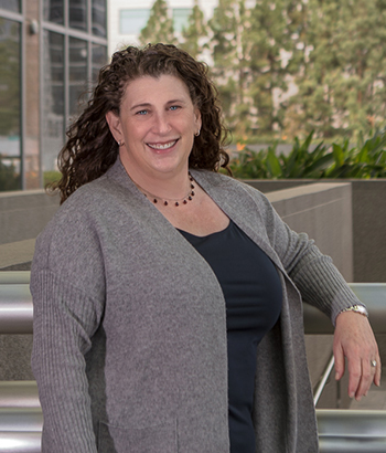 A photo of Lisa Lainer standing in front of an office building.  She is facing the camera with a smile on her face.  She is wearing a dark blue top with a grey sweater over it.