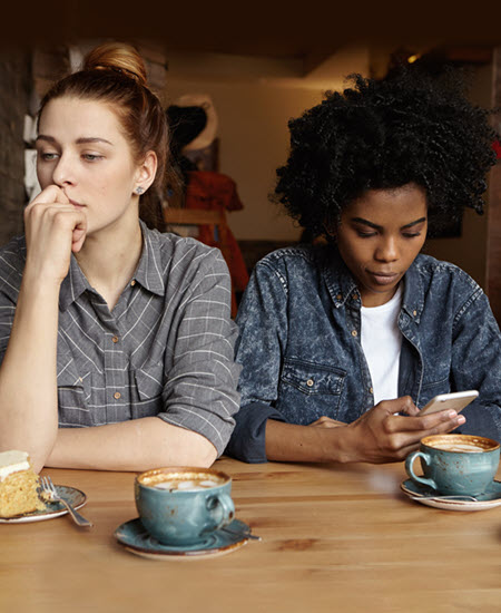 This is a photo of two women sitting next to each other. The woman on the left is white and she is wearing a grey plaid shirt.  She is looking to her right and down.  Her chin is resting on her right hand. The woman on the right is black and wearing a blue shirt over a white shirt.  She is looking down at her phone which is in her right hand.