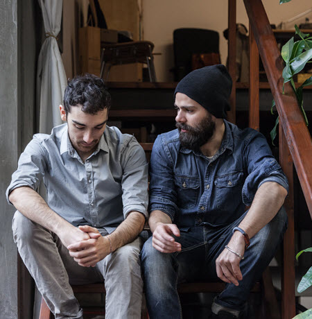 A photo of two men sitting at the bottom of a staircase.  The man on the right is wearing grey pants with a grey shirt. He is looking down at his hands which are clasp together in front of him.  The man on the right is looking at the man on the left.  He is wearing blue jeans and blue shirt and a black beanie. He has a beard.