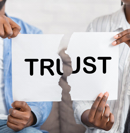 A photo of two people who are sitting on a couch.  You can not see their faces, only their bodies and their hands.  In their hands they are holding a piece of paper which has the word trust written on it.  The paper is ripped in half.