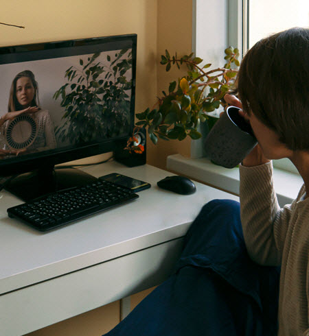 A photo of a woman looking at her computer screen.  We see her front the right and behind so we don't see her face.  The woman is sipping from a mug. On the screen is a woman holding what looks like a plate. 